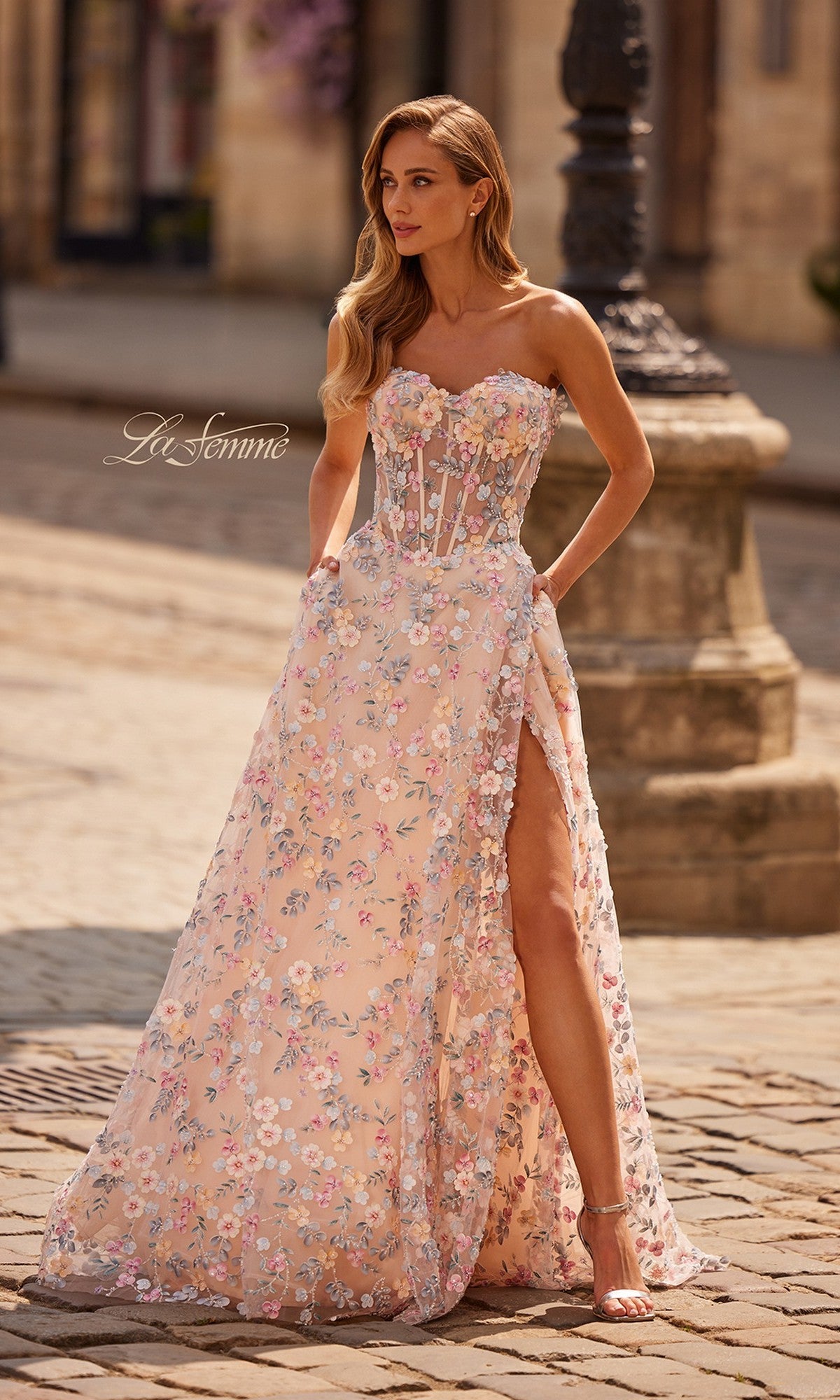 A woman outdoors on a cobblestone street wears the La Femme 33409 long prom dress in multi, featuring a strapless sweetheart neckline, floral design, thigh-high slit, and sheer bodice.